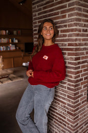 Pull doux légèrement ample bordeaux porté par une femme, vue de profil, mur en briques, jean