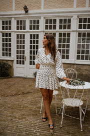 Robe cache-cœur à pois portée par une femme, silhouette entière, portes vitrées, chaise blanche