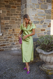Robe satinée à col plissé portée par une femme, vue de face, mur en pierre, escarpins violets