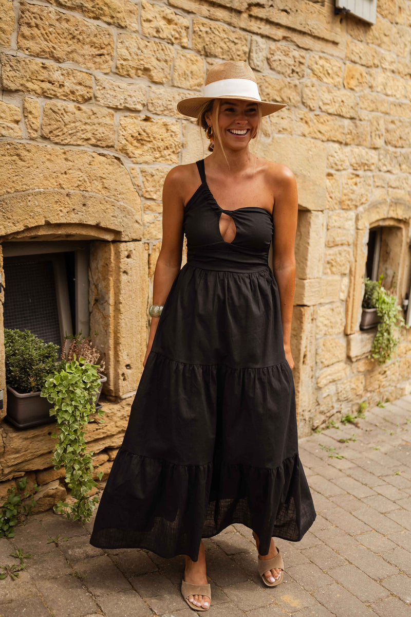 Robe longue effet lin portée par une femme, silhouette entière, façade en pierre, chapeau