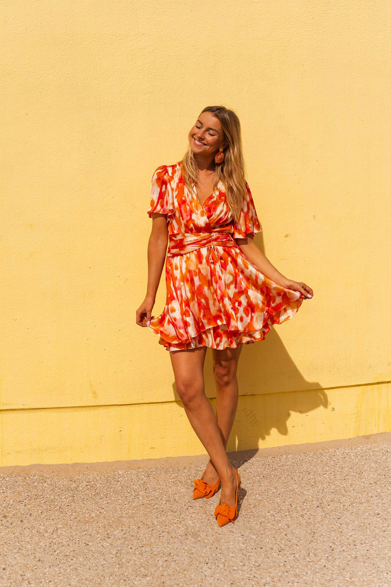 Robe cache-cœur fluide rose et orange portée par une femme, vue de face, mur jaune, escarpins orange