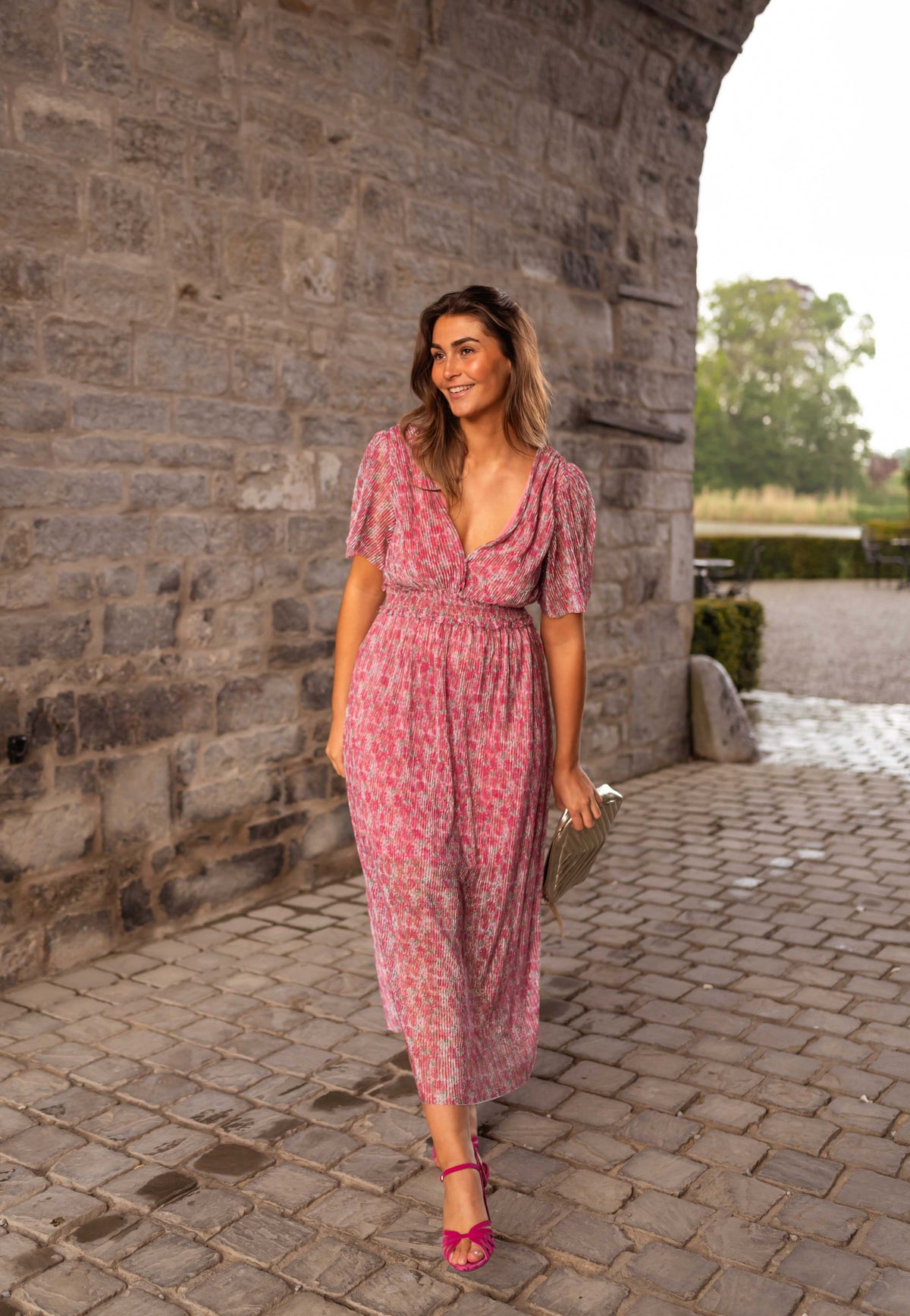 Robe plissée à col V portée par une femme, silhouette entière, mur en pierre, cour pavée