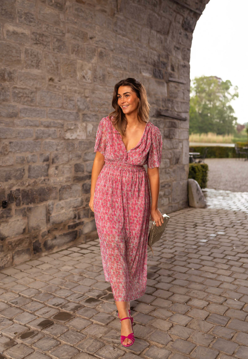 Robe plissée à col V portée par une femme, silhouette entière, mur en pierre, cour pavée