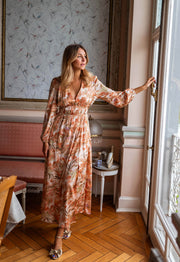 Robe longue fluide col V portée par une femme, vue de trois-quarts, fenêtre, main sur le rideau