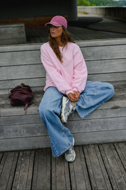 Sweat ample col rond porté par une femme, assise, casquette rose, assise sur un banc en bois
