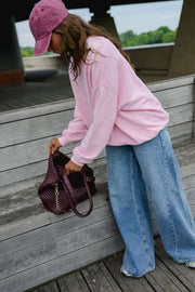 Sweat ample col rond porté par une femme, vue de trois-quarts, casquette rose, sac bordeaux, jean large, penchée