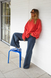 Sweat ample à col rond porté par une femme, vue de trois-quarts, mur blanc, tabouret bleu