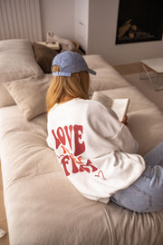 Sweat à col rond blanc porté par une femme, vue de dos, lit, casquette bleue, inscription rouge