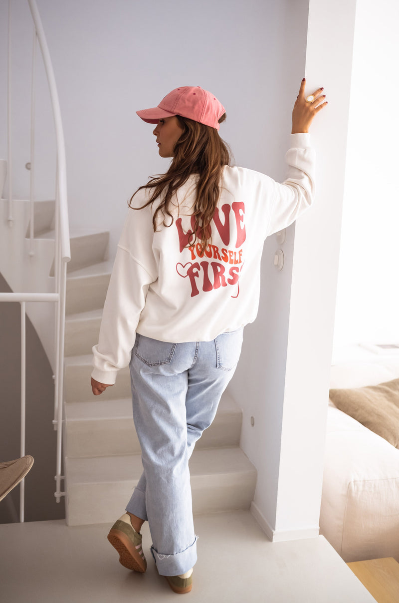 Sweat à col rond blanc porté par une femme, vue de trois-quarts, escalier blanc, casquette rose