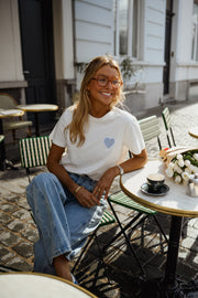 T-shirt blanc à cœur vichy bleu porté par une femme, assise, à une table, lunettes