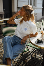 T-shirt blanc à cœur vichy bleu porté par une femme, assise, à une table, chaise verte