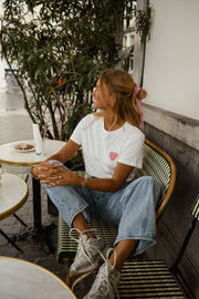 T-shirt blanc à cœur vichy rouge porté par une femme, assise, à une table, baskets blanches