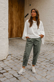 T-shirt resserré à la taille porté par une femme, vue de trois-quarts, pantalon vert-gris, devant un mur