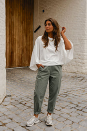 T-shirt resserré à la taille porté par une femme, silhouette entière, pantalon vert-gris, devant un mur en bois