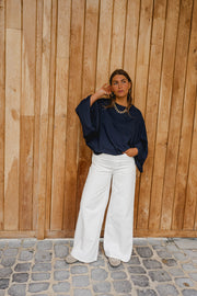 T-shirt à manches chauve-souris porté par une femme, silhouette entière, pantalon blanc, devant un mur en bois