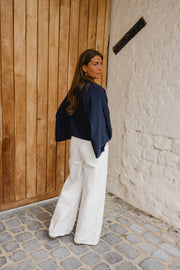 T-shirt à manches chauve-souris porté par une femme, vue de dos, pantalon blanc, devant un mur en bois