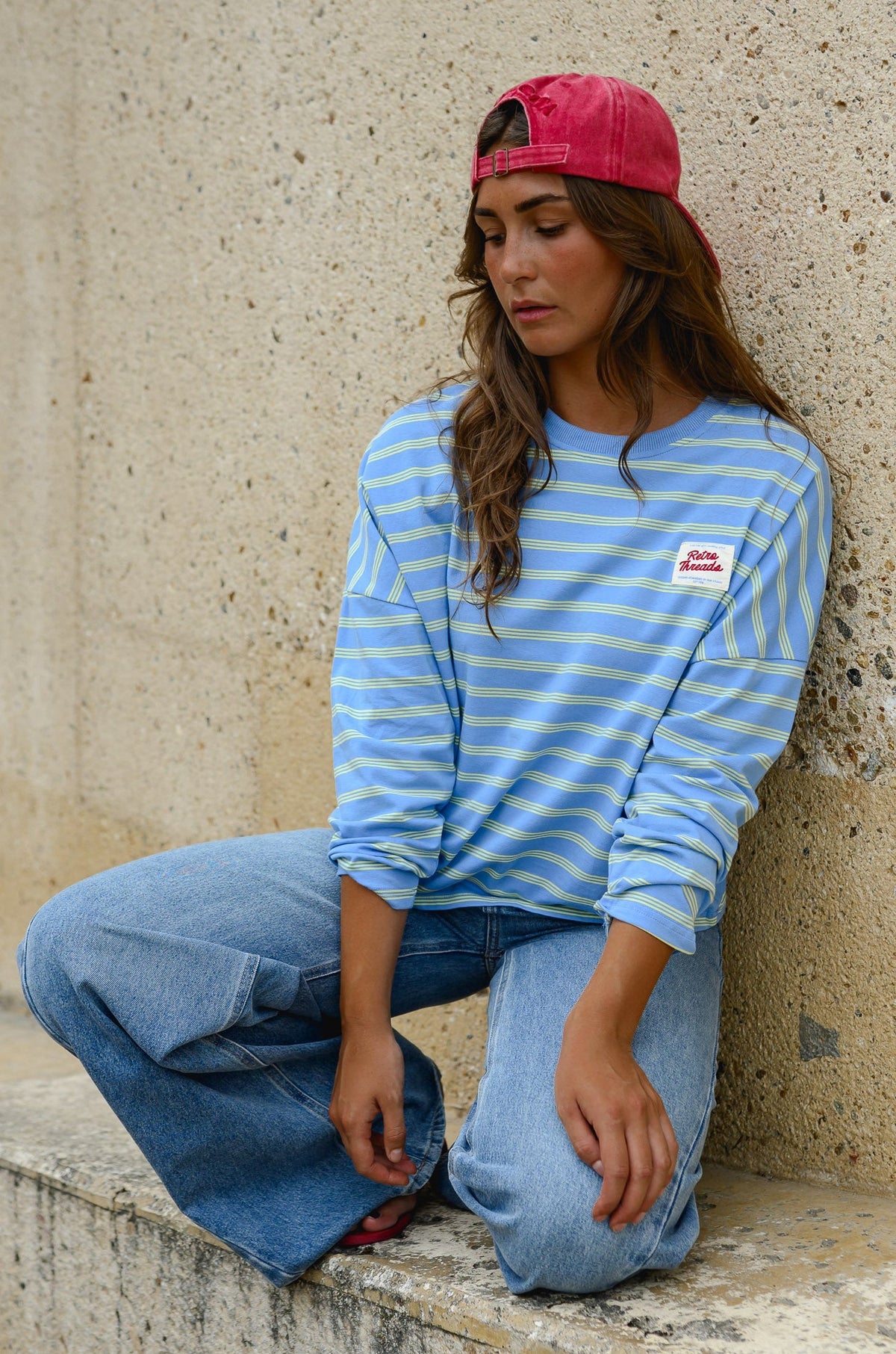 T-shirt rayé à patch porté par une femme, assise, adossée à un mur, casquette rouge