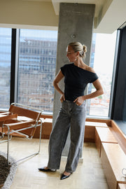 T-shirt moulant asymétrique noir porté par une femme, silhouette entière, près d'une fenêtre