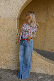 T-shirt fleuri asymétrique moulant porté par une femme, silhouette entière, vue de face, près d'un mur courbe