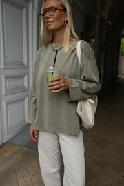 T-shirt loose à manches longues porté par une femme, silhouette entière, lunettes de soleil, boisson verte, sac blanc, devant une porte grise