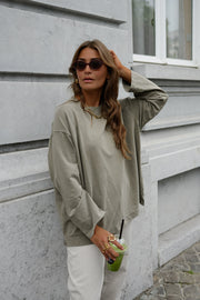 T-shirt loose à manches longues porté par une femme, vue de trois-quarts, lunettes de soleil, main sur la tête, mur gris