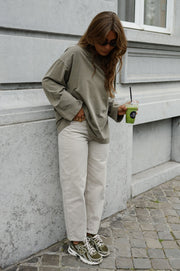 T-shirt loose à manches longues porté par une femme, vue de trois-quarts, adossée à un mur, boisson verte, baskets