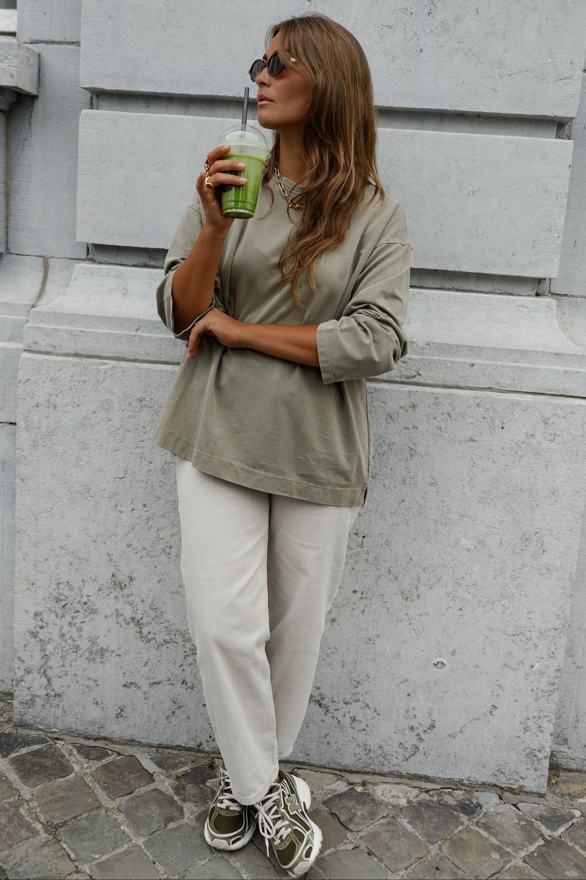 T-shirt loose à manches longues porté par une femme, vue de trois-quarts, pantalon beige, boisson verte à la main, adossée à un mur