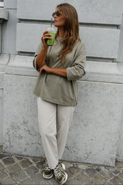 T-shirt loose à manches longues porté par une femme, vue de trois-quarts, pantalon beige, boisson verte à la main, adossée à un mur