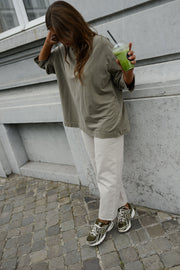 T-shirt loose à manches longues porté par une femme, vue de dos, cheveux longs, adossée au mur, boisson verte