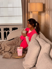 Gilet mouton sans manches rose porté par une femme, assise, sur un canapé, plaid, gobelet rose