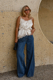 Top satiné à fines bretelles porté par une femme, silhouette entière, jean large bleu, mur en pierre
