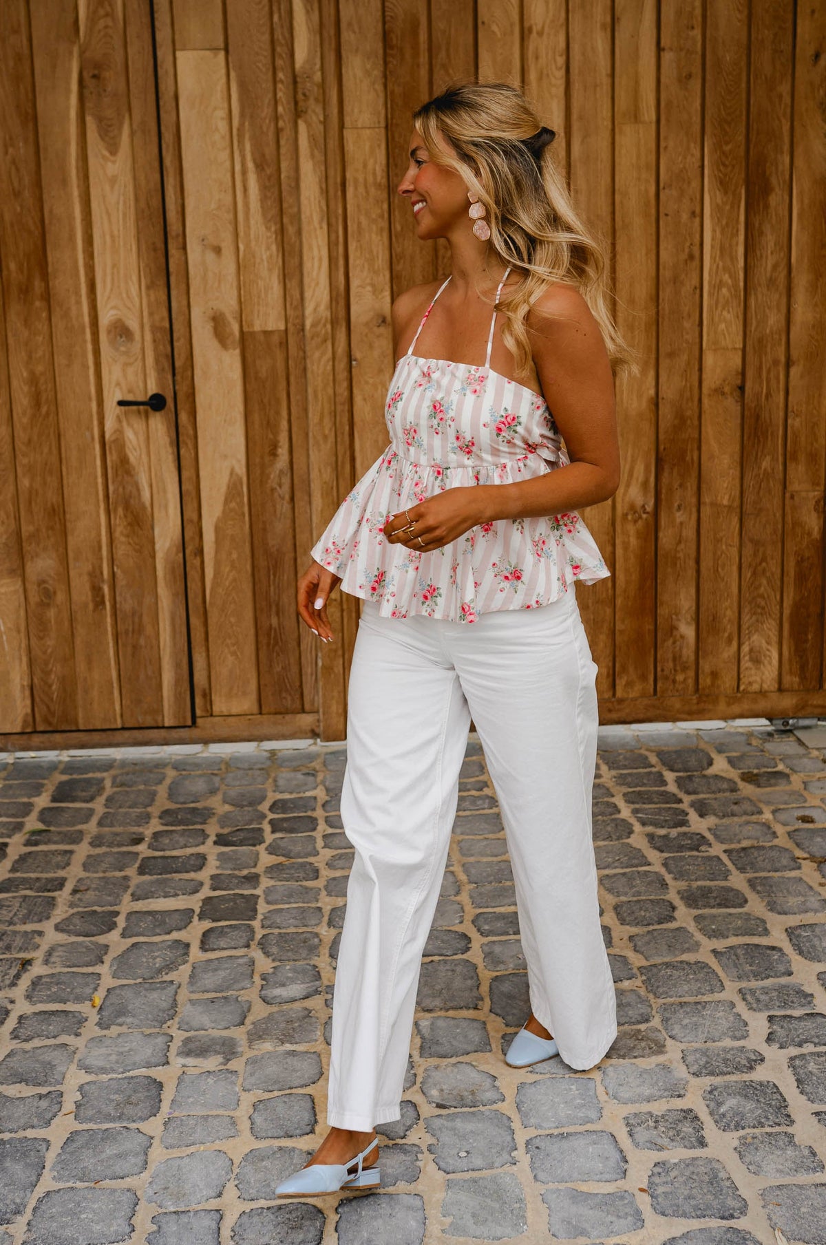 Top fleuri dos ouvert noué porté par une femme, silhouette entière, pantalon blanc, porte en bois