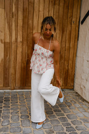 Top fleuri dos ouvert noué porté par une femme, silhouette entière, pantalon blanc, porte en bois, de côté