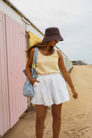 Top fluide effet lin jaune porté par une femme, silhouette entière, short blanc, chapeau, sac bleu