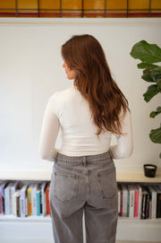 T-shirt moulant col rond écru porté par une femme, vue de dos, jean gris, bibliothèque