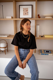 T-shirt à manches courtes noir porté par une femme, assise, étagères en bois, jean bleu