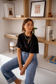 T-shirt à manches courtes noir porté par une femme, assise, étagères en bois, sourire