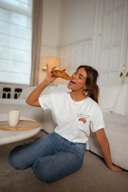 T-shirt à logo croissant blanc porté par une femme, assise, assise au sol, croissant à la main