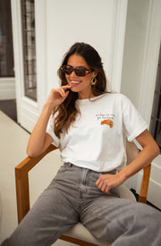 T-shirt à logo croissant blanc porté par une femme, assise, lunettes de soleil, chaise en bois