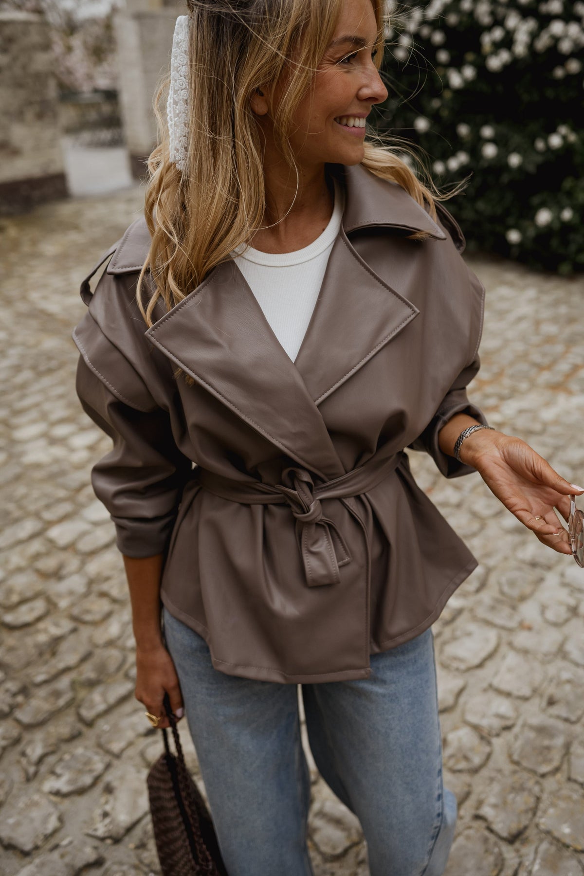 Veste effet cuir ceinturée portée par une femme, vue de trois-quarts, jean, sourire