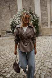 Veste effet cuir ceinturée portée par une femme, silhouette entière, sac à la main, jean, buissons fleuris