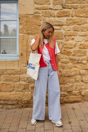 Veste sans manches boutonnée rouge portée par une femme, vue de trois-quarts, fenêtre, sac en toile