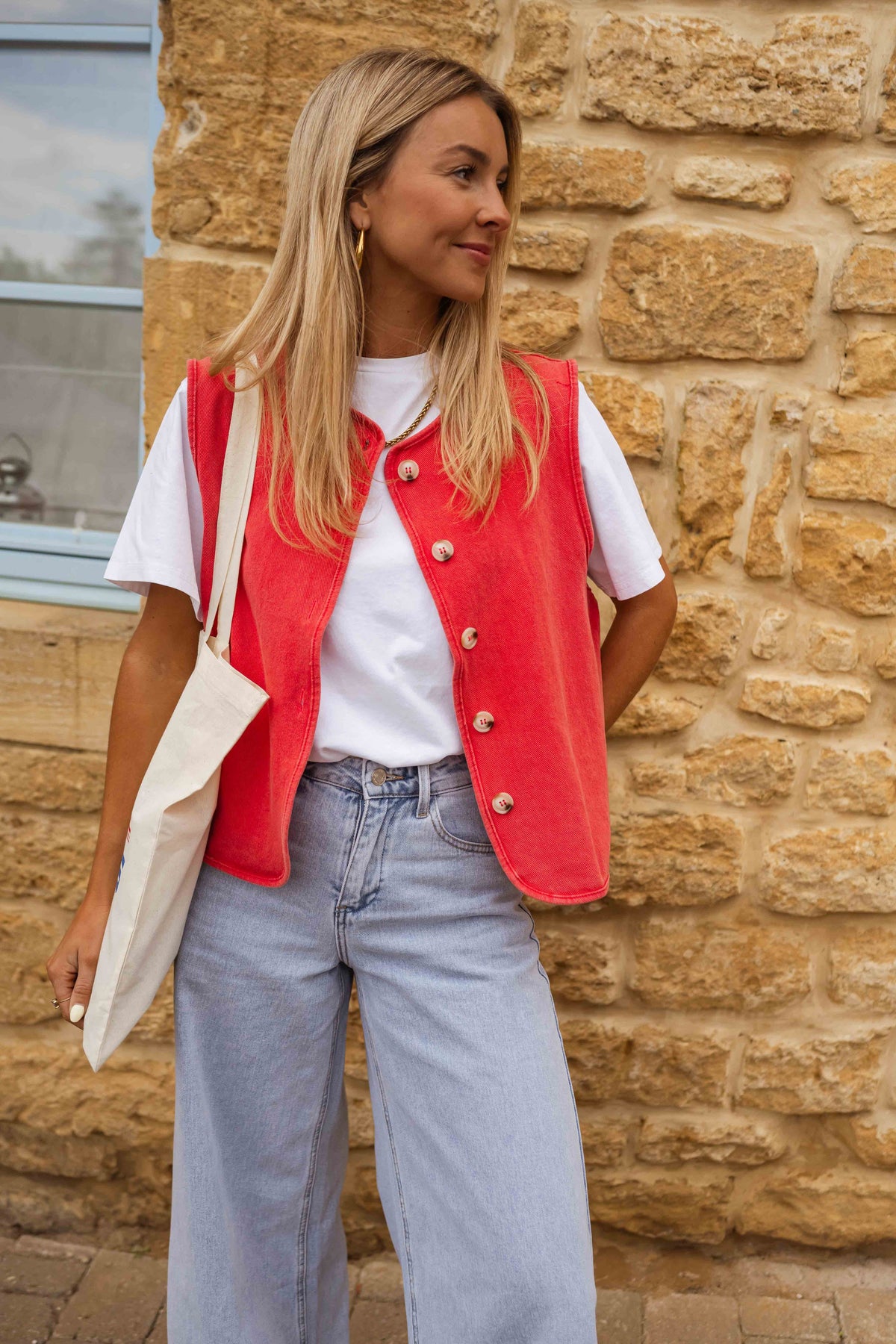Veste sans manches boutonnée rouge portée par une femme, vue de trois-quarts, mur en pierre, sac en toile