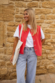 Veste sans manches boutonnée rouge portée par une femme, vue de face, mur en pierre, sac en toile