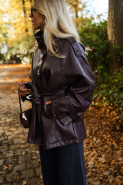 Veste en simili cuir ceinturée bordeaux portée par une femme, vue de trois-quarts, feuilles d'automne, sac à main
