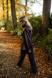 Veste en simili cuir ceinturée bordeaux portée par une femme, vue de profil, allée pavée, jean bleu