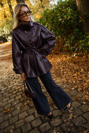 Veste en simili cuir ceinturée bordeaux portée par une femme, vue de trois-quarts, lunettes de soleil, sac noir