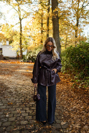 Veste en simili cuir ceinturée bordeaux portée par une femme, vue de face, sous-bois, ceinture nouée