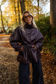 Veste en simili cuir ceinturée bordeaux portée par une femme, vue de trois-quarts, feuilles dorées, main dans la poche