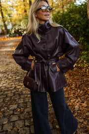 Veste en simili cuir ceinturée bordeaux portée par une femme, vue de face, allée pavée, lunettes de soleil